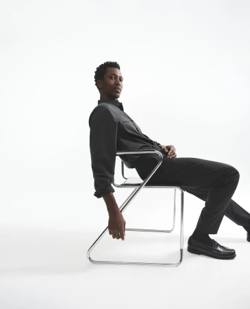 Man seated on a modern chrome cantilever chair against a plain white studio background wearing dark shirt and trousers.
