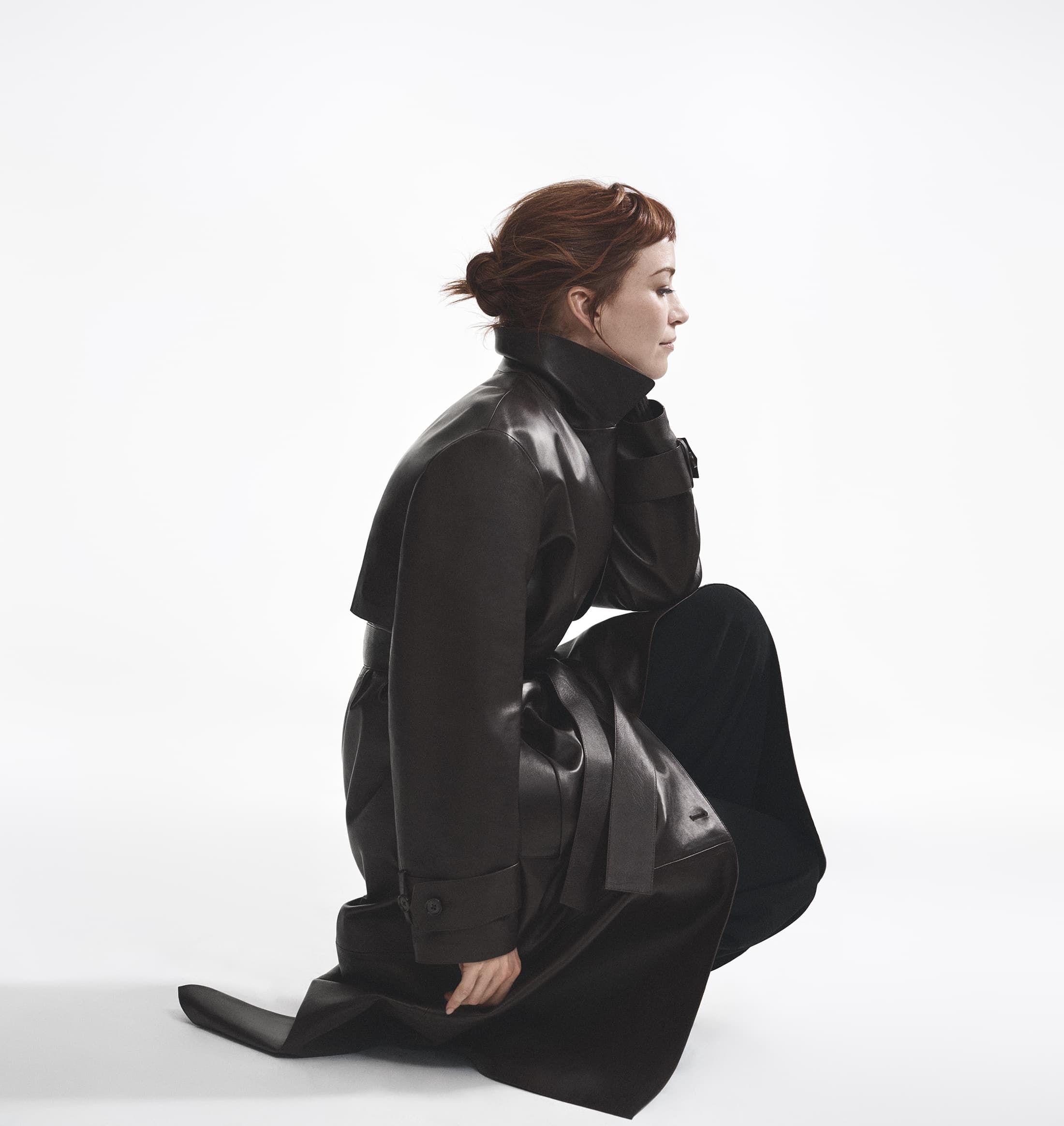 Woman kneeling in profile wearing a dark leather trench coat against a white background