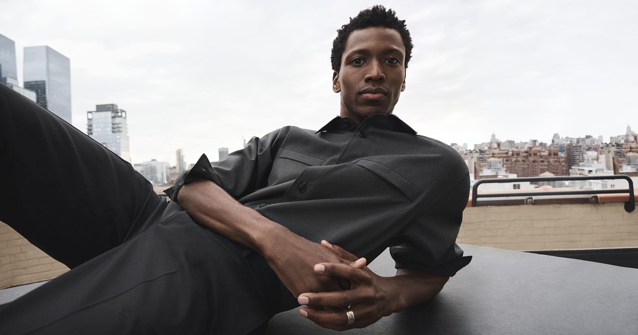 Black man reclining on a rooftop terrace wearing a dark grey shirt and trousers against a city skyline.