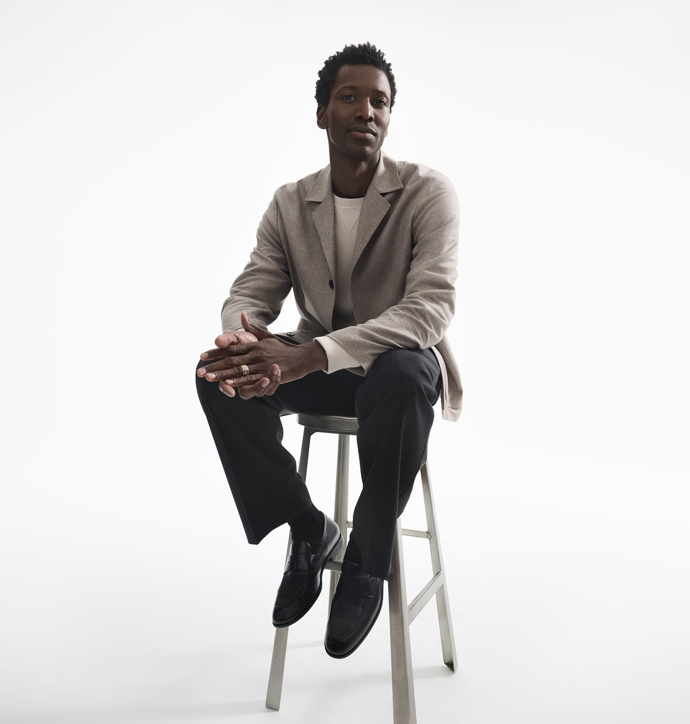 Black man in a beige blazer and dark trousers seated on a metal stool against a white background.