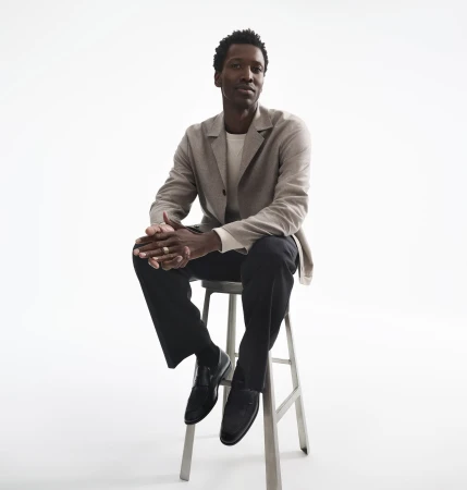Black man in a beige blazer and dark trousers seated on a metal stool against a white background.