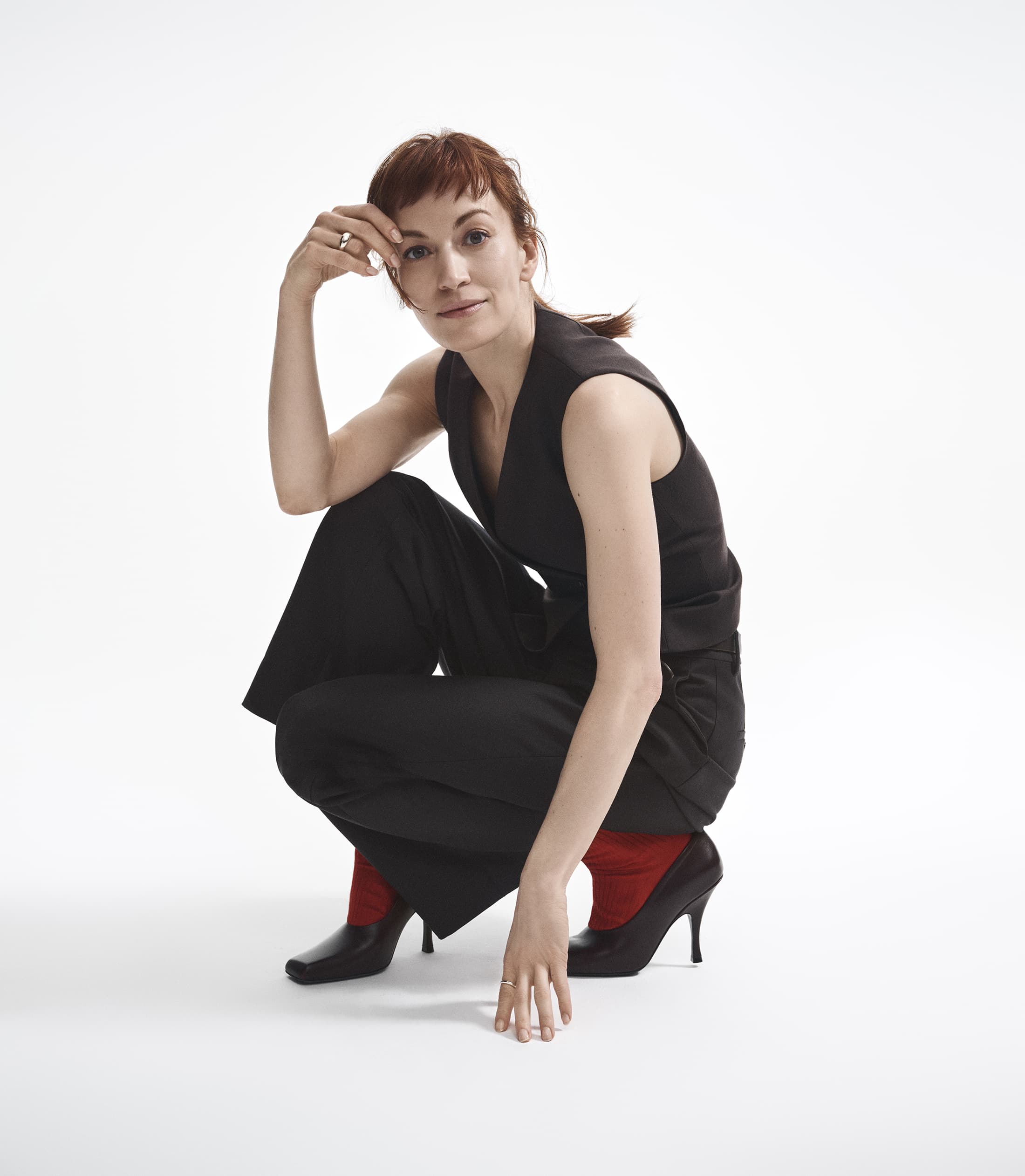 Britt Lower in black sleeveless top and trousers crouching against a white backdrop for Theory