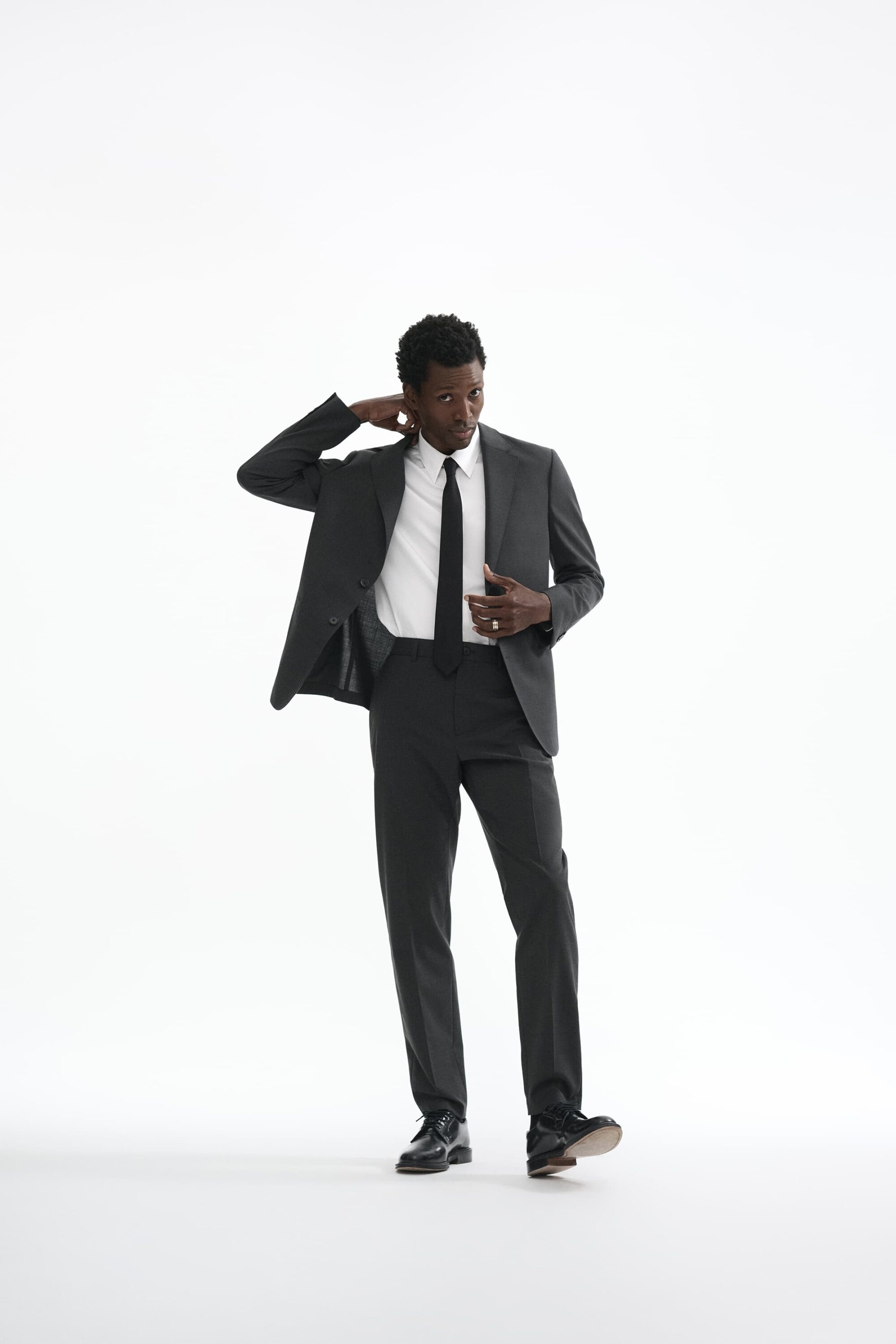 Man wearing a charcoal grey suit and white shirt with a black tie standing against a white seamless background.
