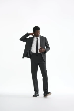 Man wearing a charcoal grey suit and white shirt with a black tie standing against a white seamless background.