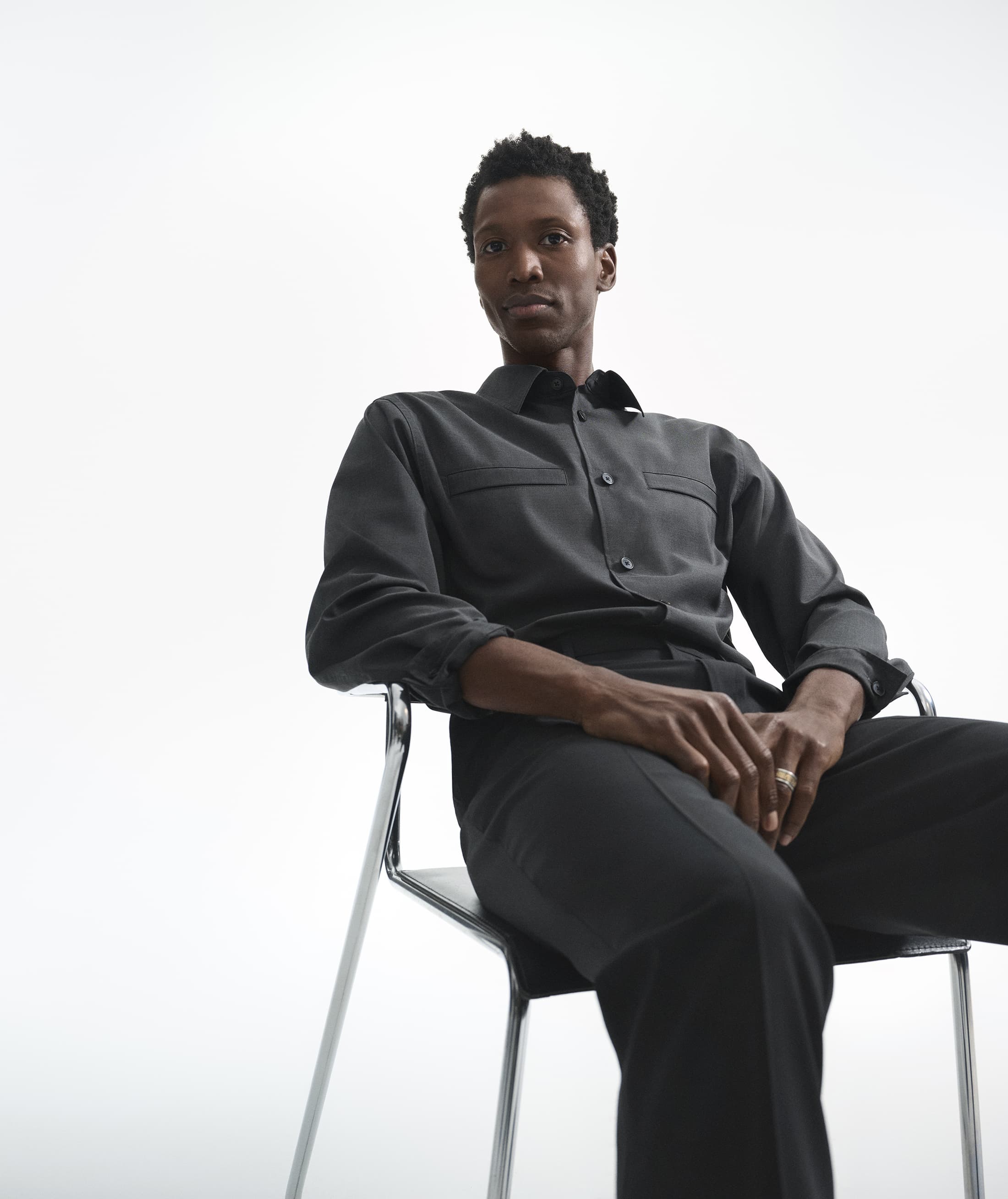 A Black man seated in a modern chrome and black chair against a plain white background, wearing a dark grey button-down shirt and trousers.