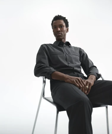 A Black man seated in a modern chrome and black chair against a plain white background, wearing a dark grey button-down shirt and trousers.