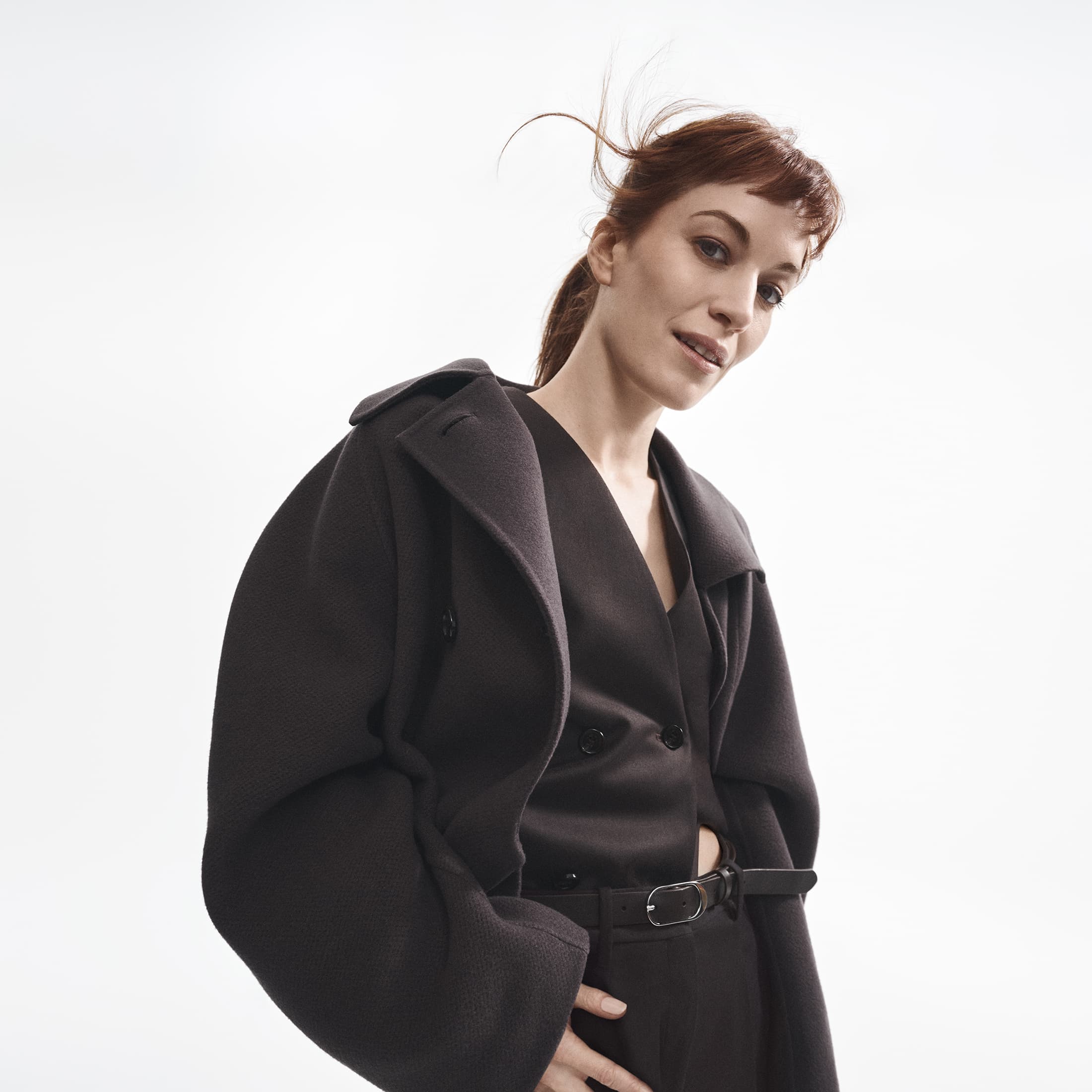 Woman with reddish brown hair wearing a dark oversized coat against a white background