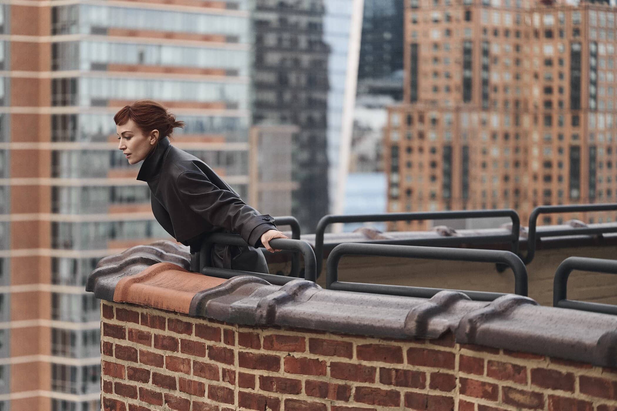 Britt Lower on a brick rooftop leaning against a railing overlooking city buildings for Theory fall 2025 campaign