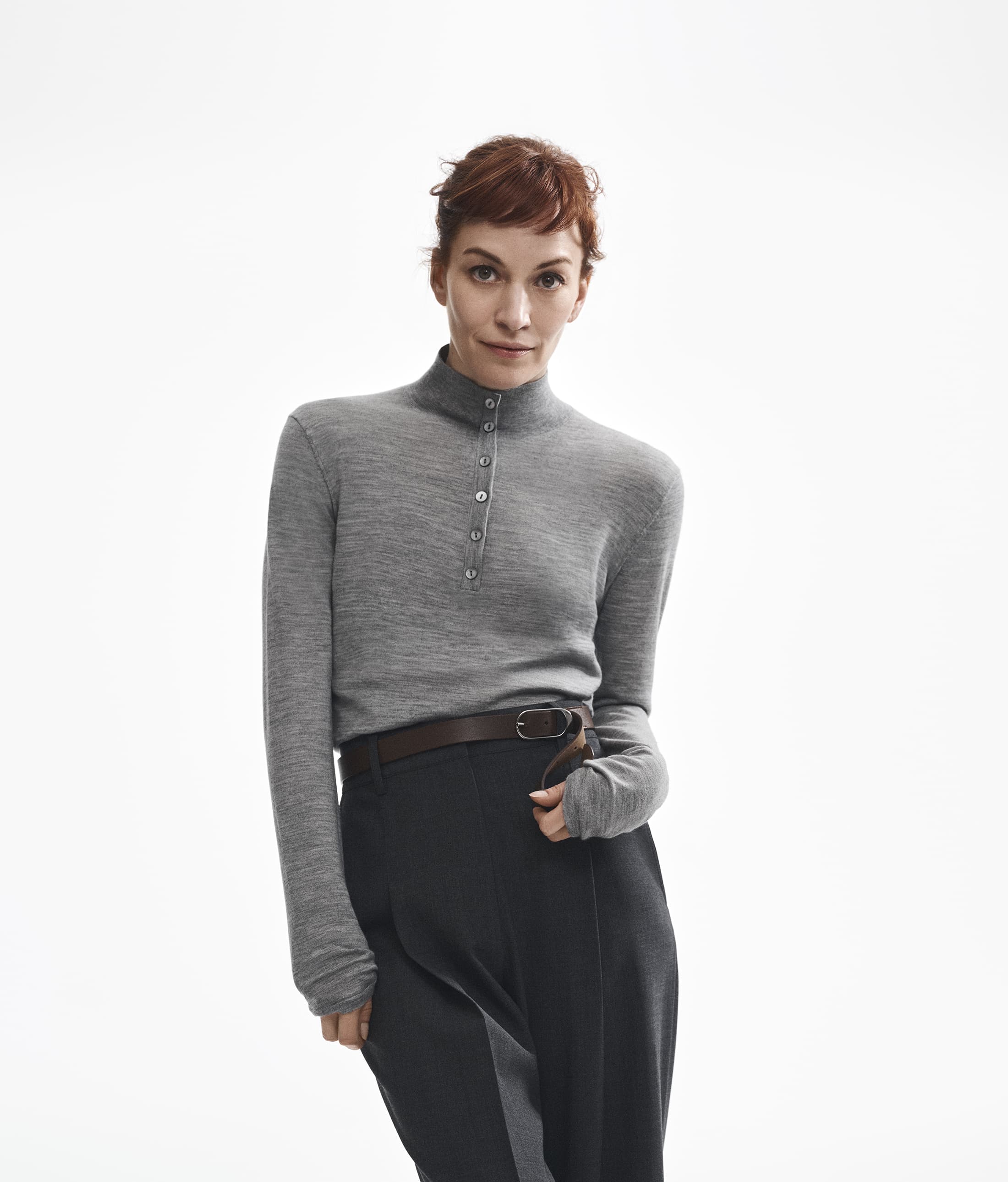 Britt Lower wearing a grey buttoned turtleneck and dark trousers against a white background for Theory.