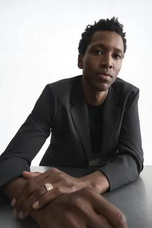 Portrait of a Black man in a dark blazer leaning forward with clasped hands against a bright white background.