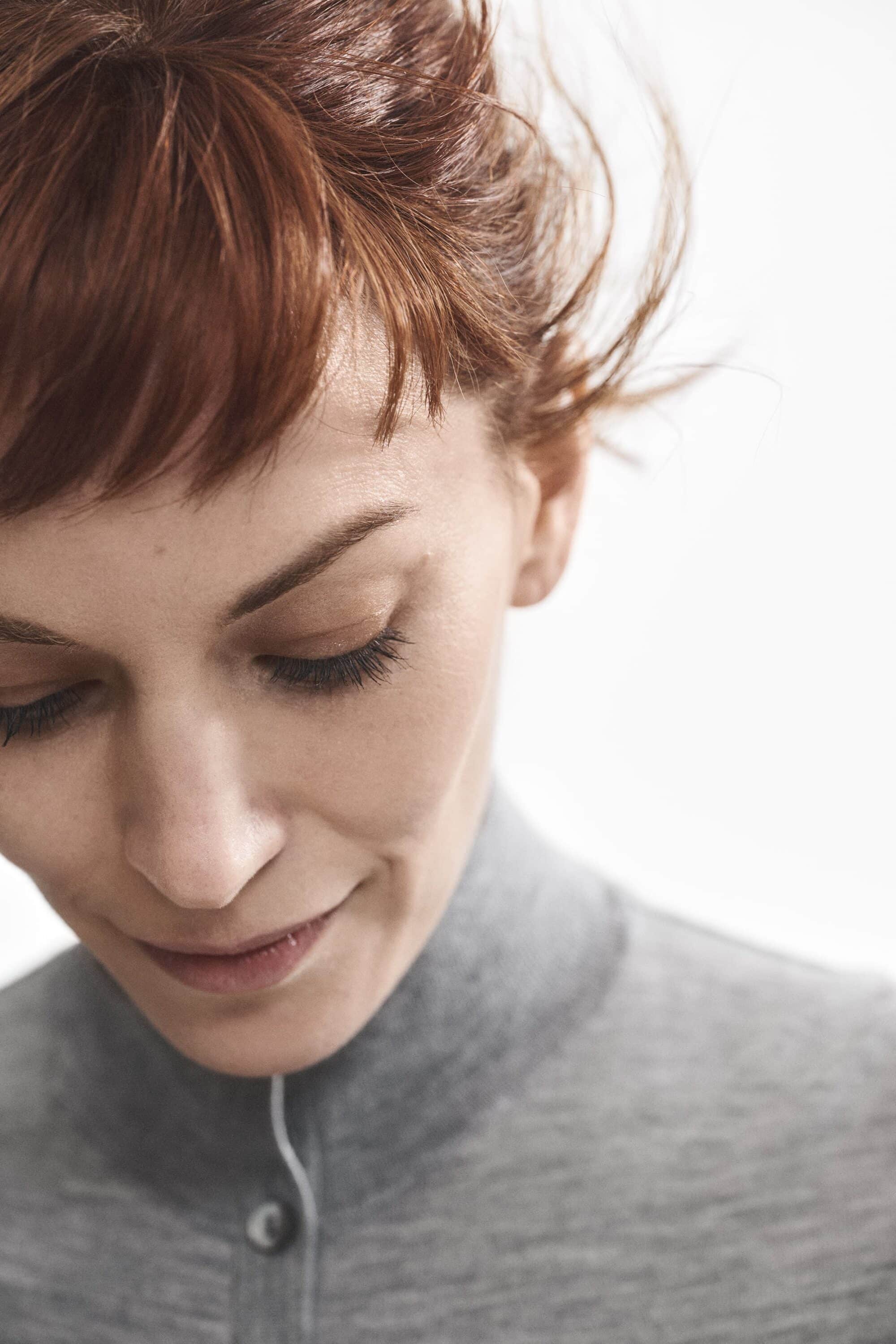 Close-up portrait of a woman with auburn hair wearing a grey turtleneck sweater against a bright white background