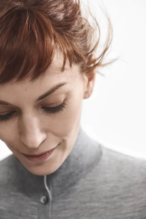 Close-up portrait of a woman with auburn hair wearing a grey turtleneck sweater against a bright white background