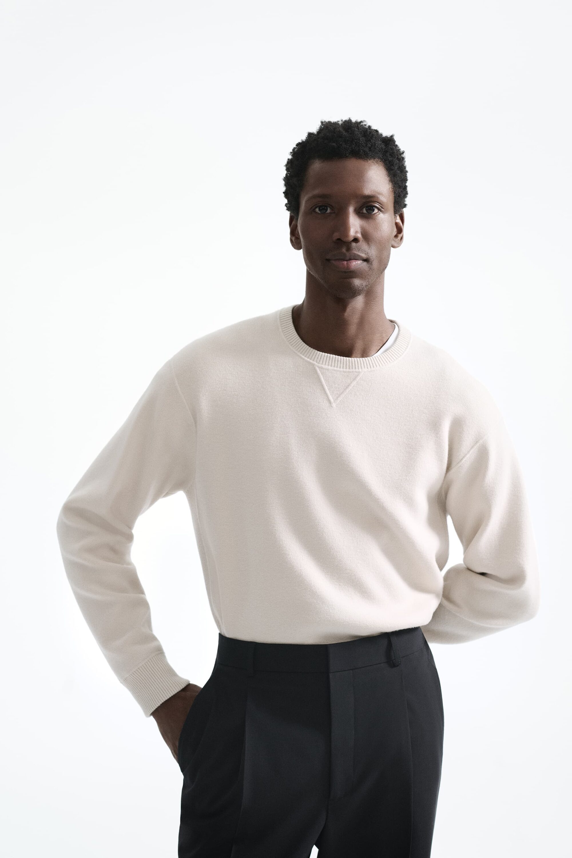 Black man wearing a cream crewneck sweater and dark trousers against a white background