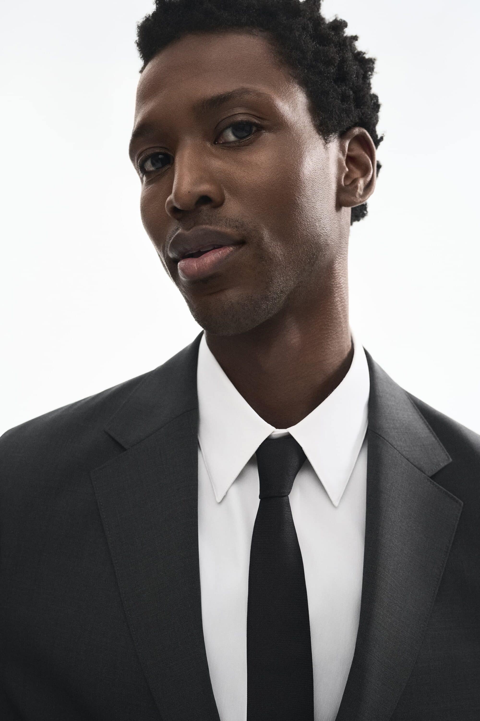 Portrait of a Black man wearing a dark suit jacket, white shirt, and black tie against a white background