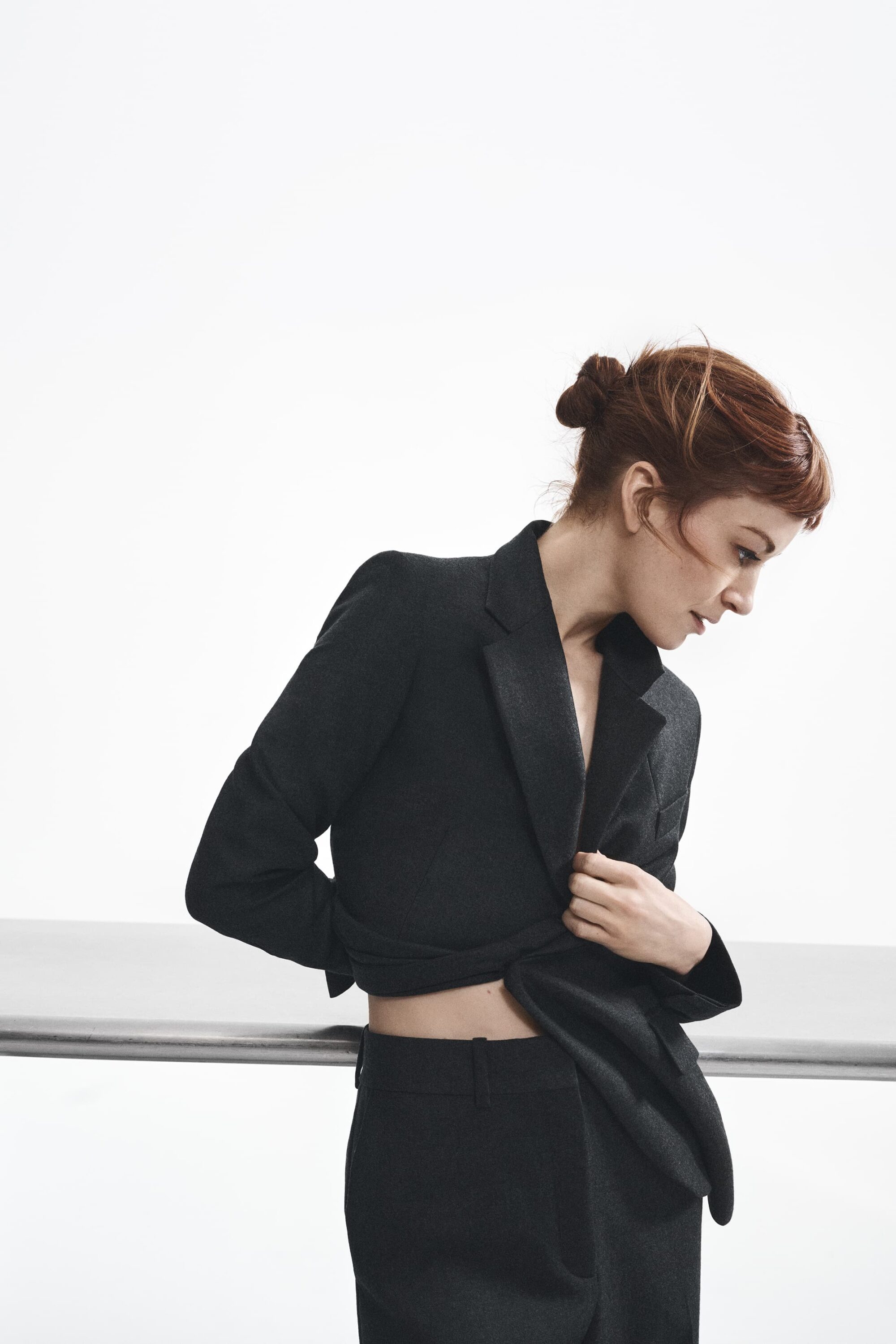 Woman in a black tailored suit leaning against a railing against a plain white studio background