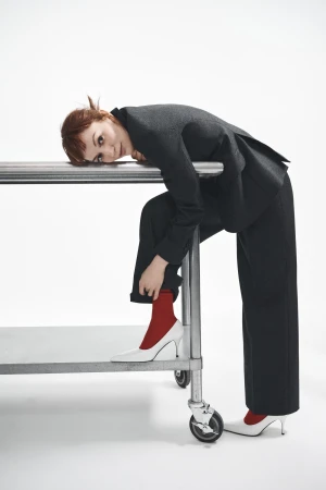 Woman in dark suit leaning on a metal utility cart wearing white heels and red socks against a white studio background