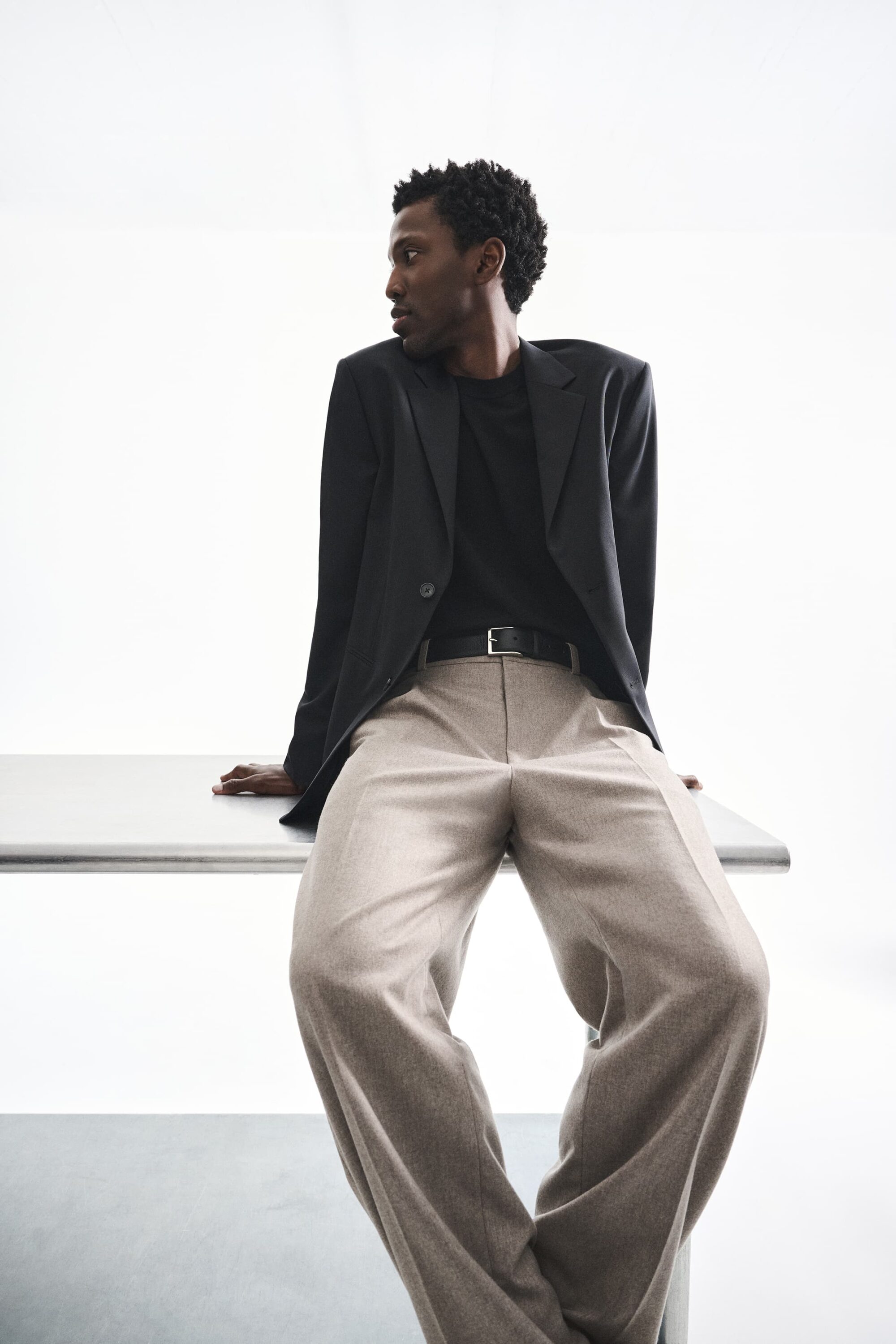 Black man seated on a metal table wearing a dark blazer, black t-shirt, and beige trousers against a white seamless backdrop.