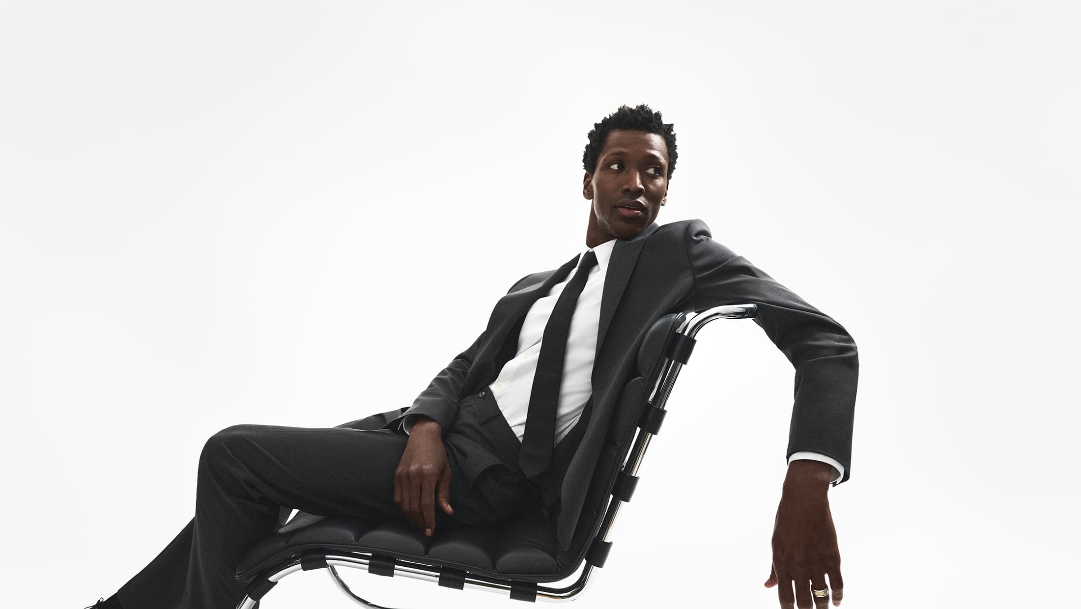 Man wearing a dark suit and tie seated on a modern black leather chair against a bright white background.