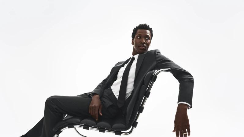 Man wearing a dark suit and tie seated on a modern black leather chair against a bright white background.