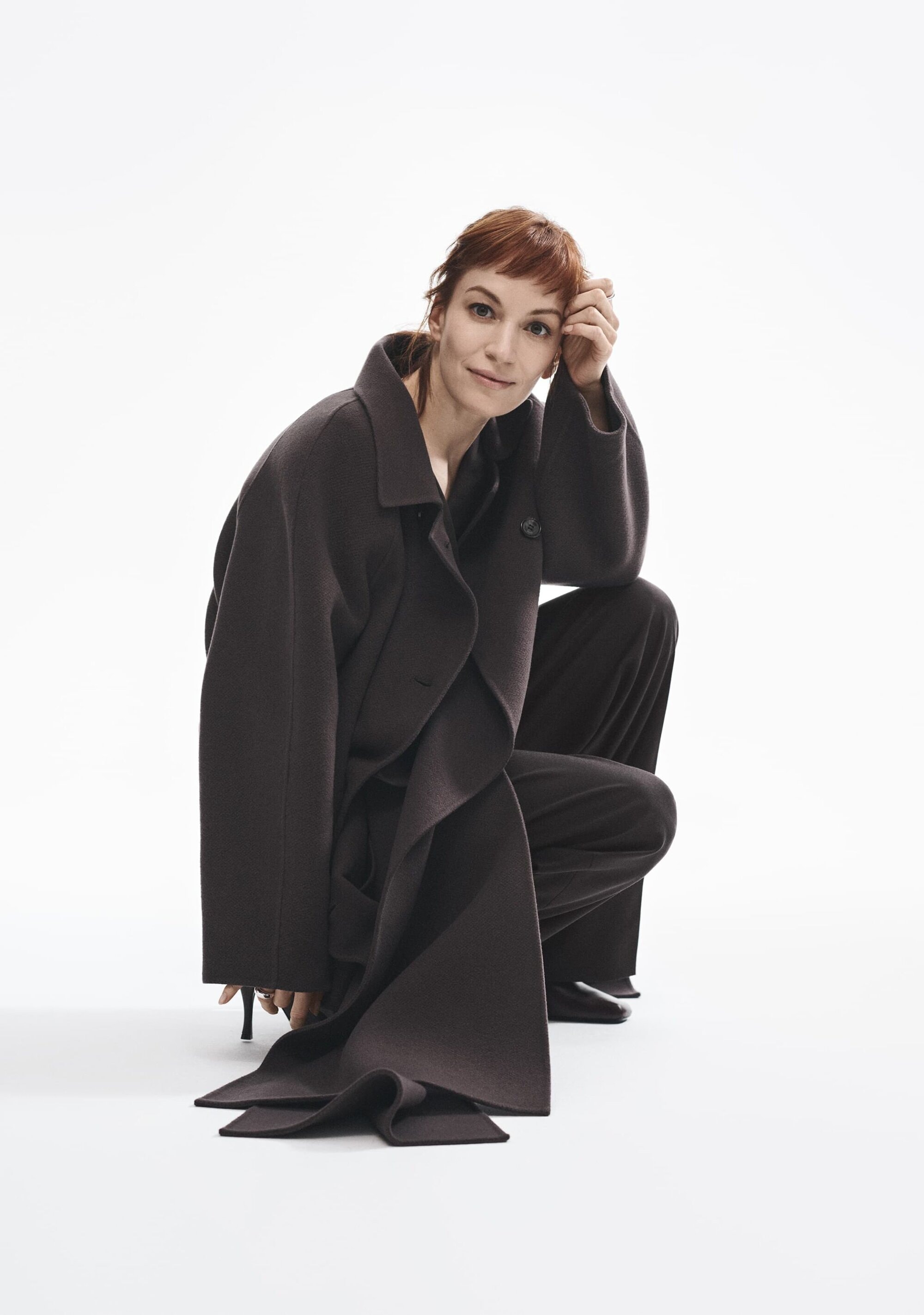 Woman with red hair wearing a dark oversized coat and trousers squatting against a plain white studio background.