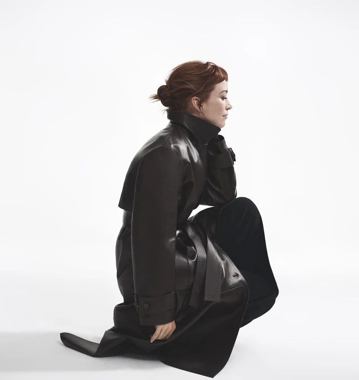 Woman kneeling in profile wearing a dark leather trench coat against a white background
