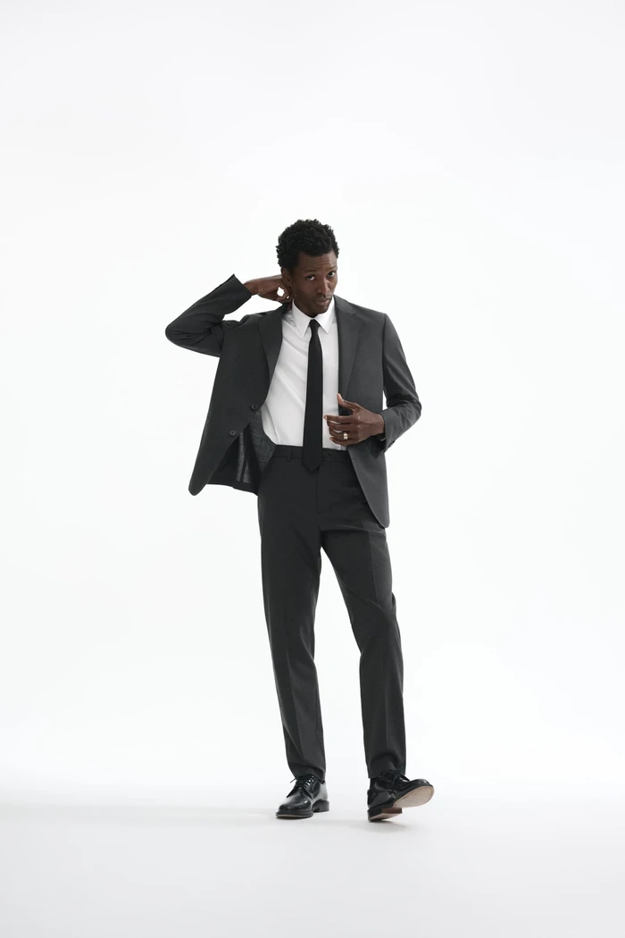 Man wearing a charcoal grey suit and white shirt with a black tie standing against a white seamless background.