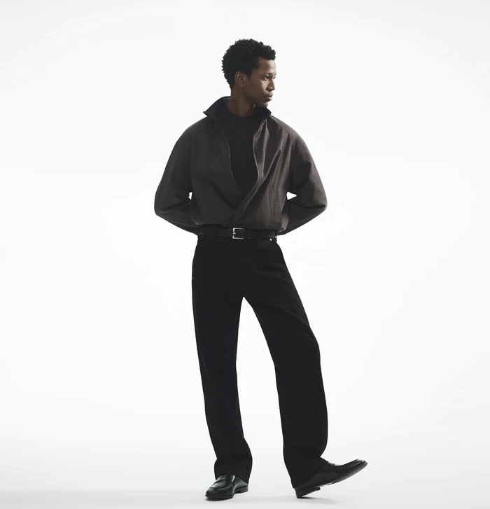 Man wearing a dark jacket and black trousers against a white seamless background for Theory fall 2025 campaign.