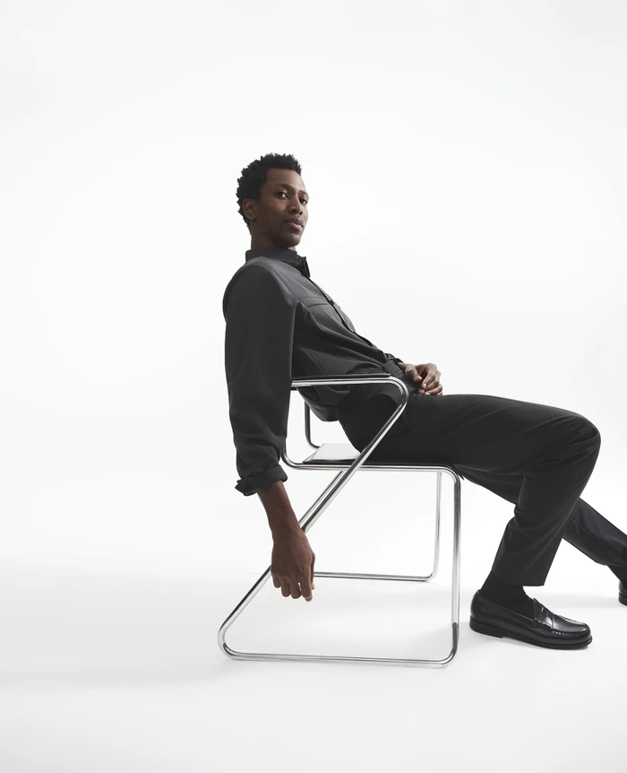 Man seated on a modern chrome cantilever chair against a plain white studio background wearing dark shirt and trousers.