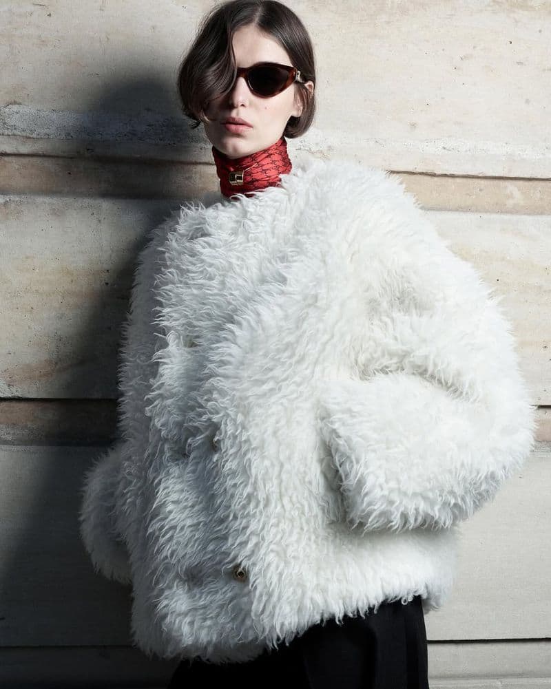 Model wearing a fluffy white textured coat and dark sunglasses standing against a stone wall for Celine