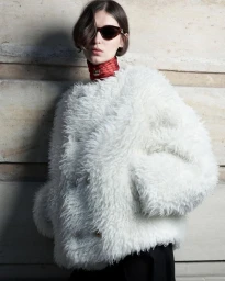 Model wearing a fluffy white textured coat and dark sunglasses standing against a stone wall for Celine