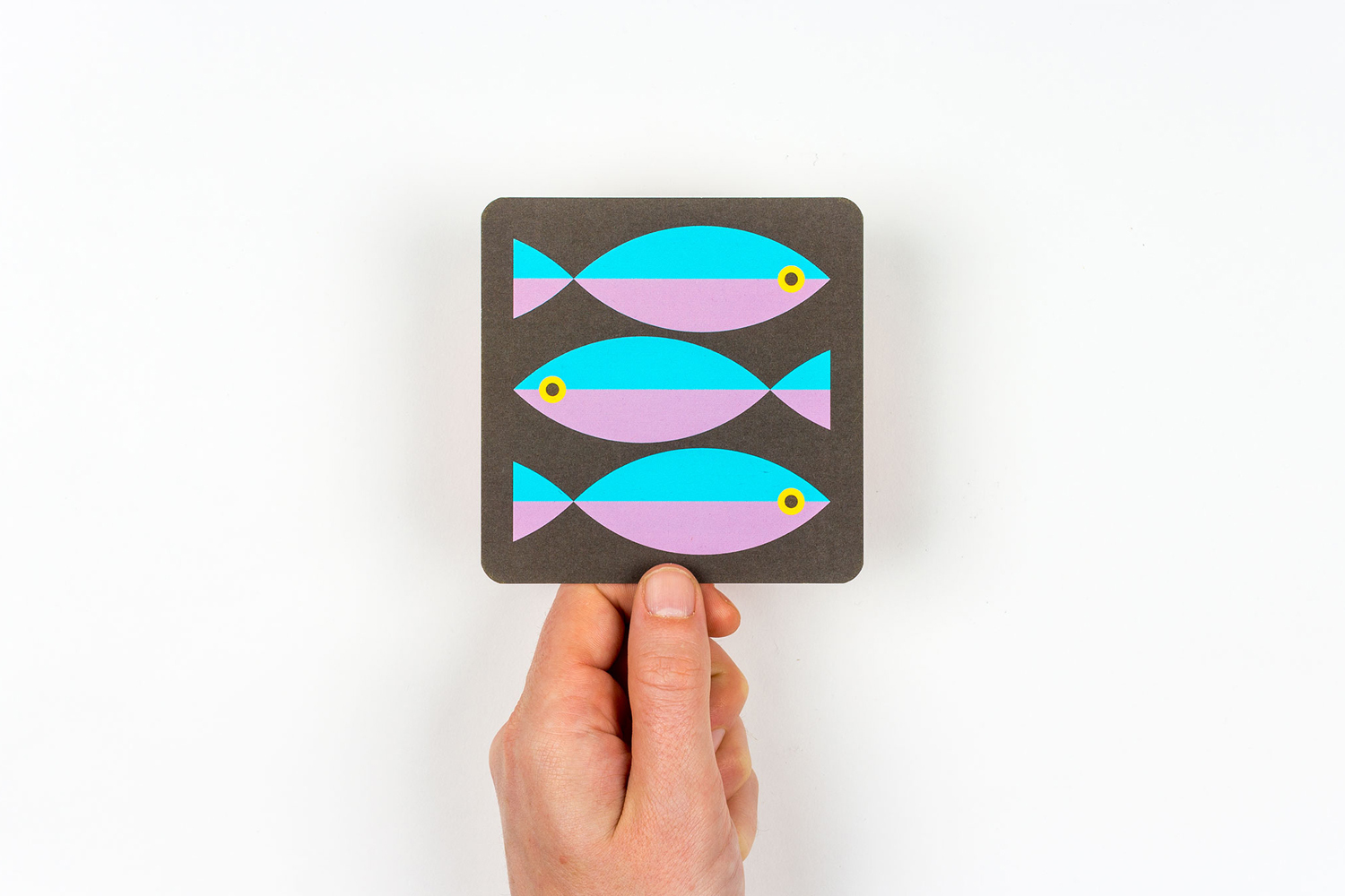 A hand holding a dark square card featuring three colorful graphic fish illustrations against a plain white background.