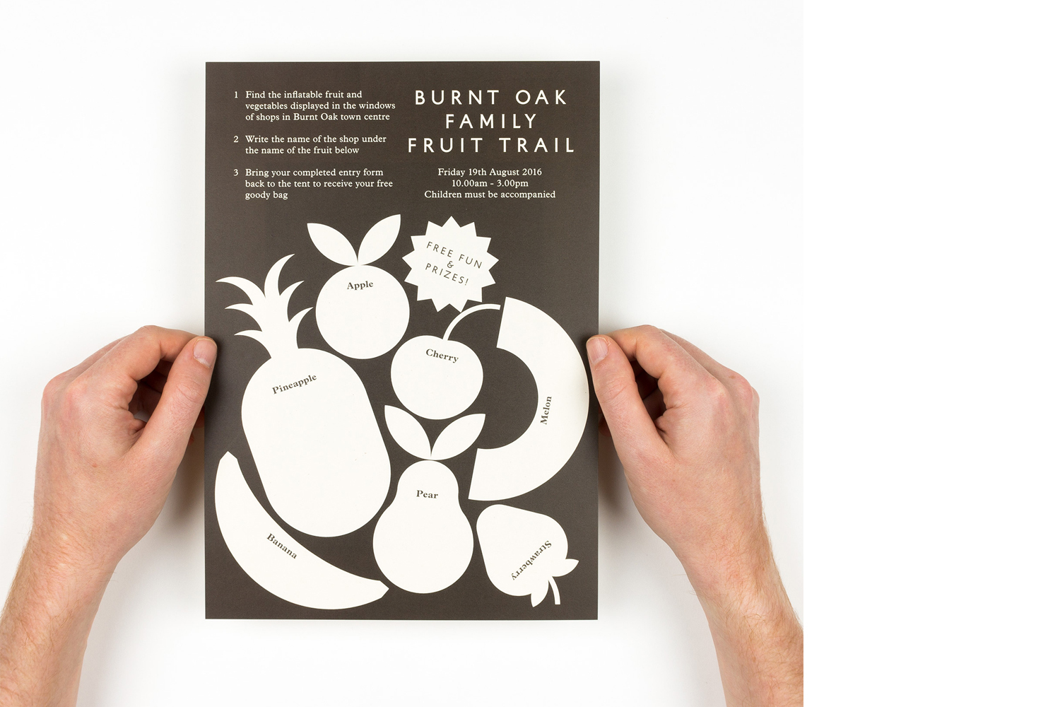 Two hands holding a dark brown and white printed flyer for the Burnt Oak Family Fruit Trail against a plain white background.