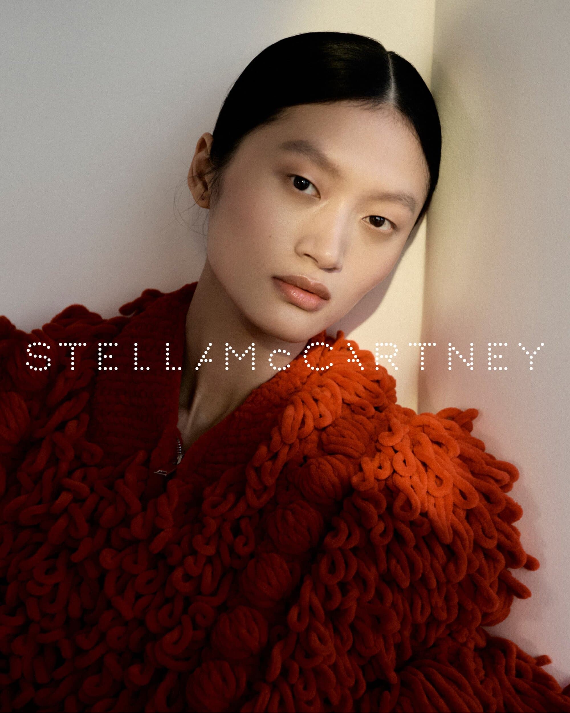 A model with slicked-back dark hair wearing a textured red knit sweater leans against a pale studio wall.