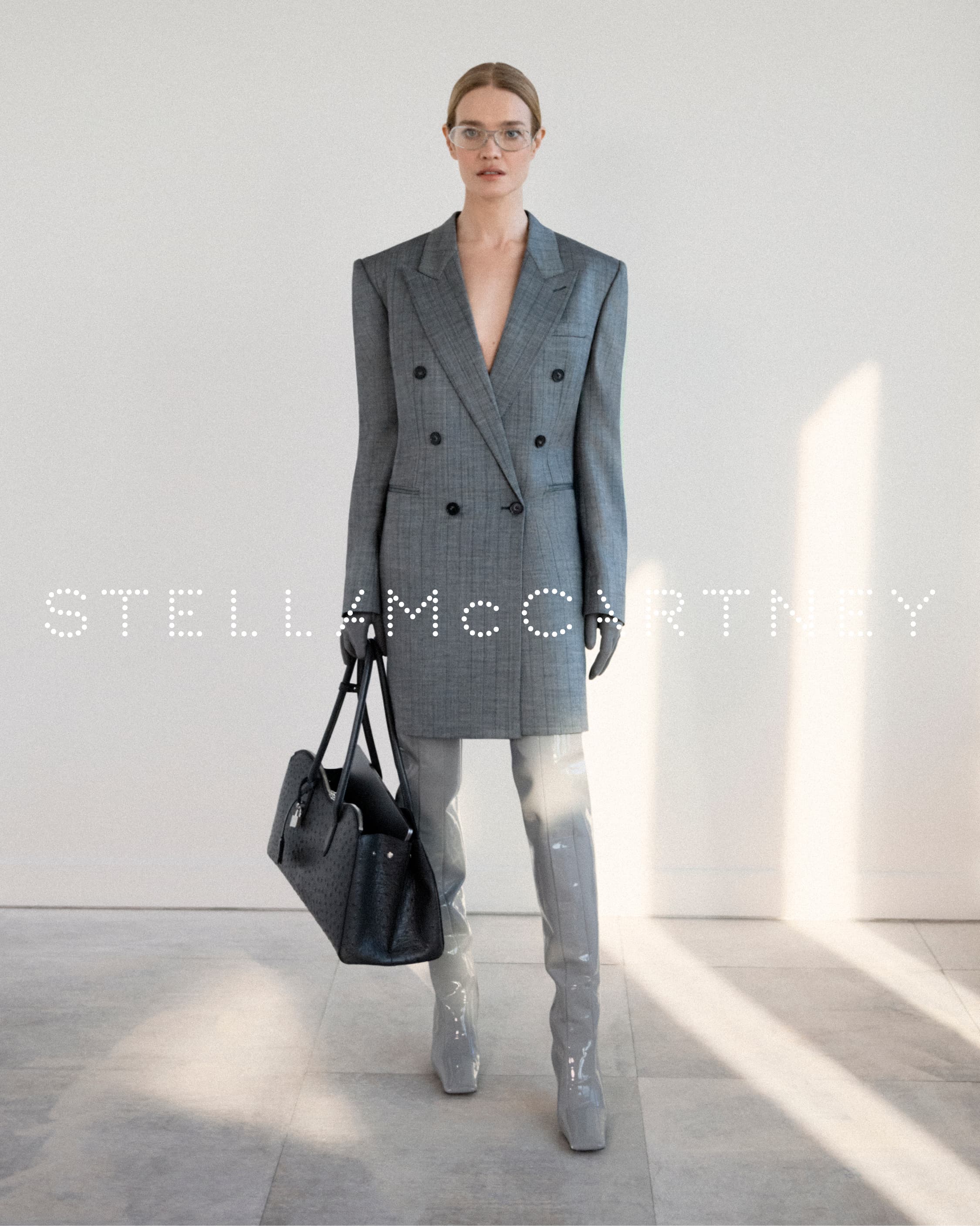 Model wearing a grey double-breasted blazer dress and patent thigh-high boots, holding a black handbag against a light wall.