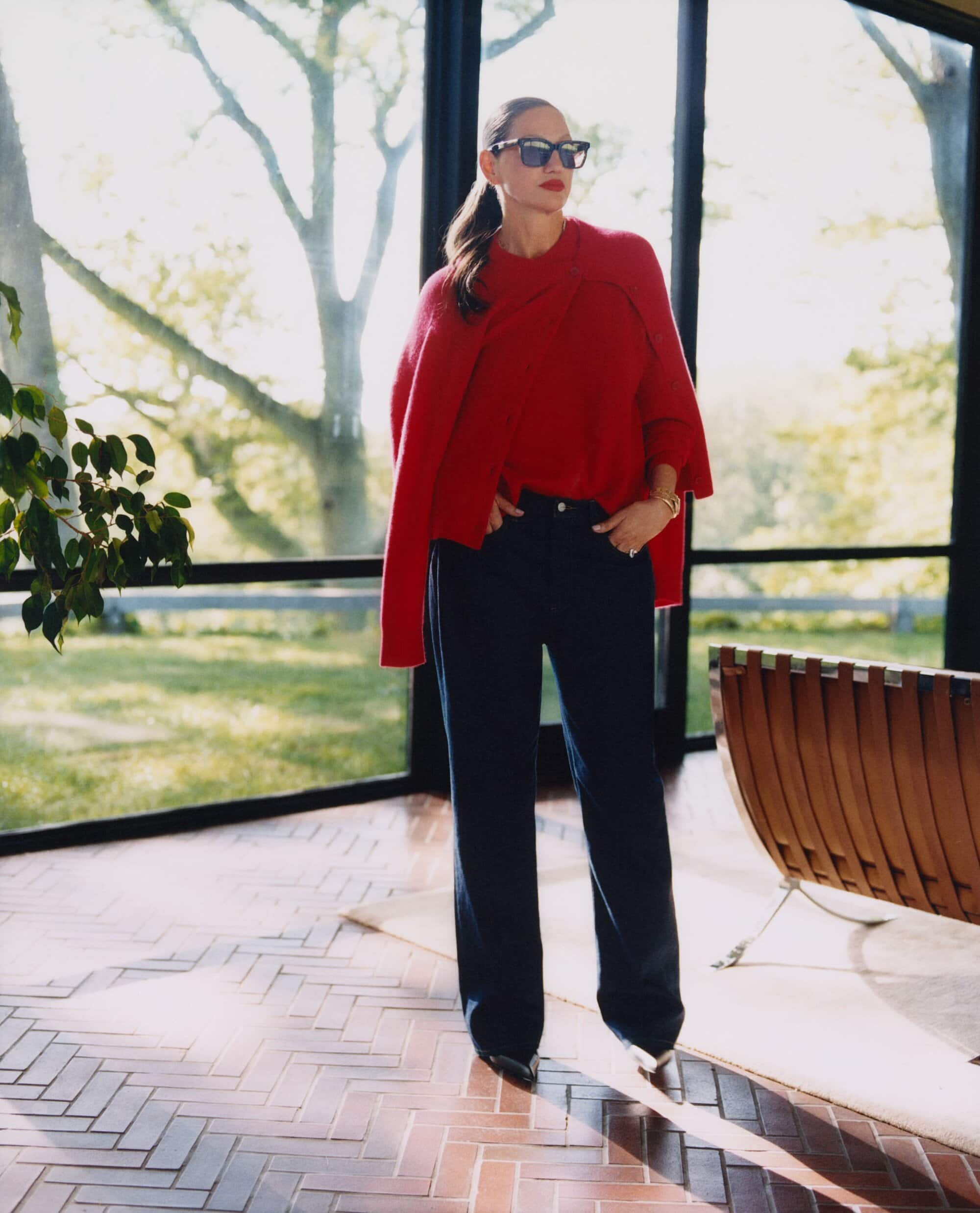 Jenna Lyons wearing red sweater and dark trousers standing indoors by large windows for Aritzia Fall 2025 Ad Campaign