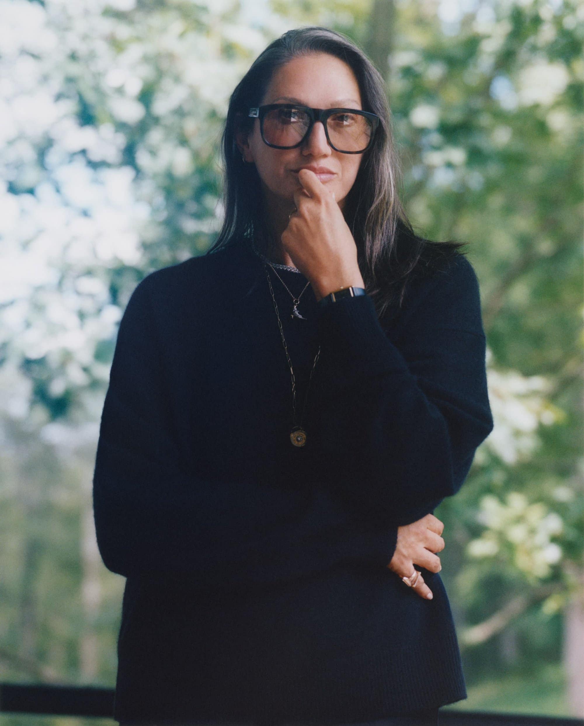 Jenna Lyons wearing black sweater and bold glasses with hand near chin outdoors for Aritzia Fall 2025 ad campaign.