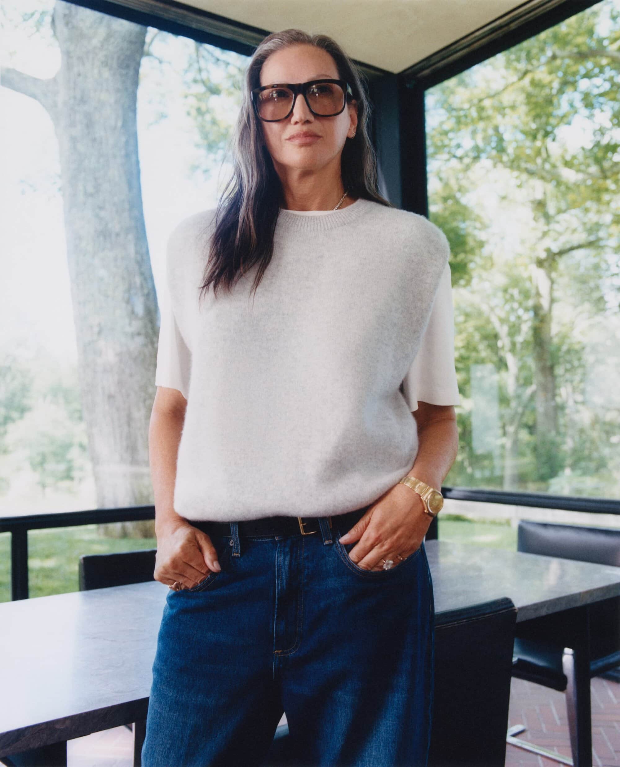 Jenna Lyons wearing oversized tinted glasses, a light grey knit vest, and dark blue jeans indoors near a large window