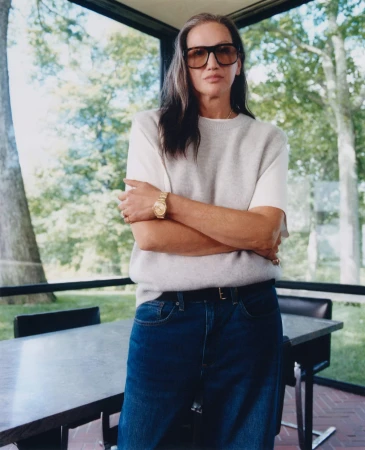 Jenna Lyons with dark sunglasses wearing a grey knit vest and blue jeans in an interior space