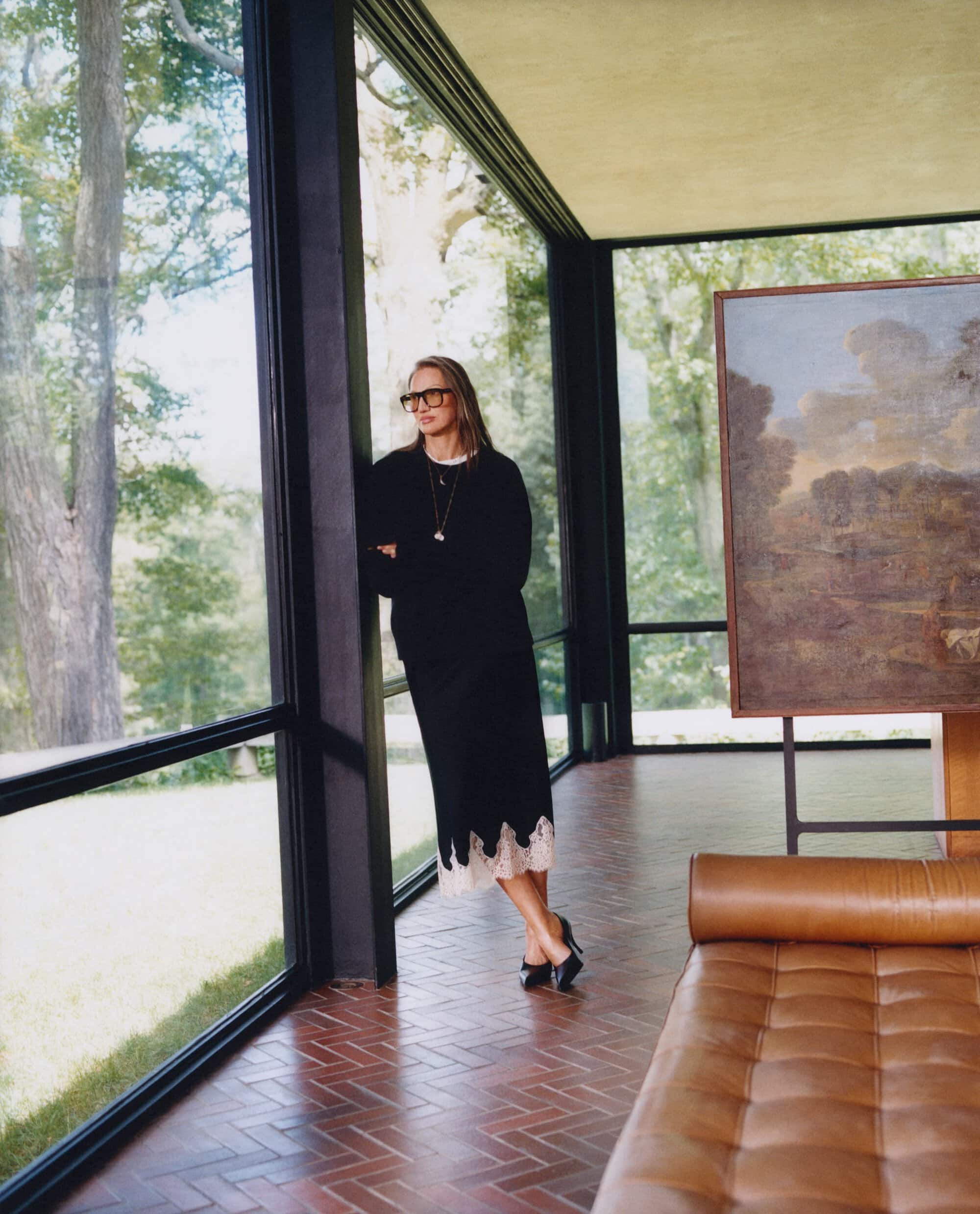 Jenna Lyons leaning against a black frame window wearing a black sweater and skirt with a tan leather daybed in foreground.