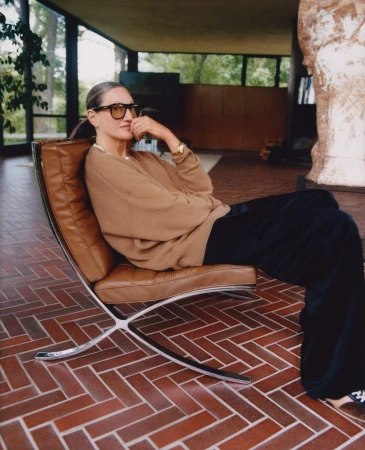 Jenna Lyons seated in a modern leather lounge chair wearing a brown sweater and dark trousers inside a room with red herringbone tile flooring.