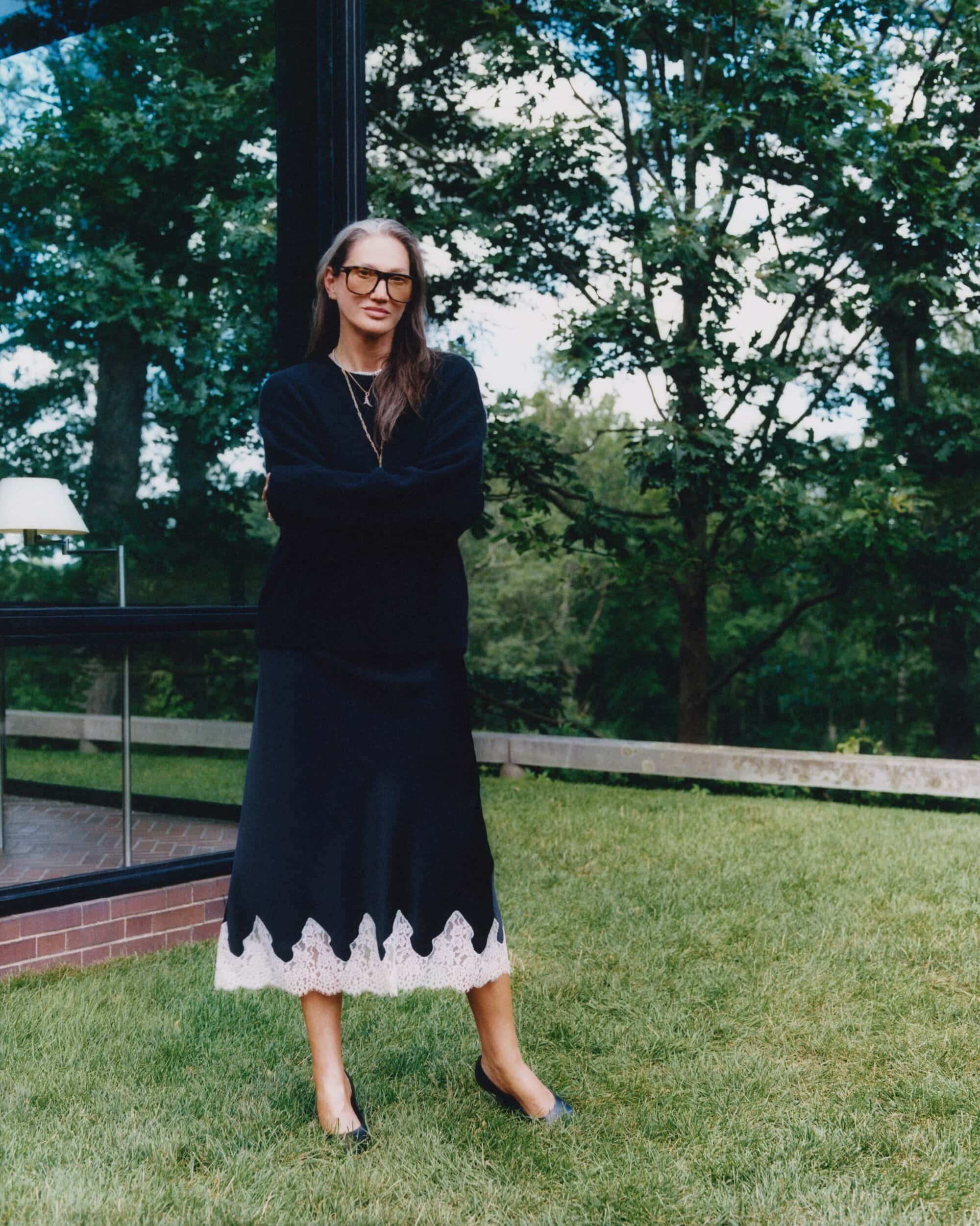 Jenna Lyons wearing black sweater and skirt with lace hem standing outdoors on grass for Aritzia Fall 2025 campaign