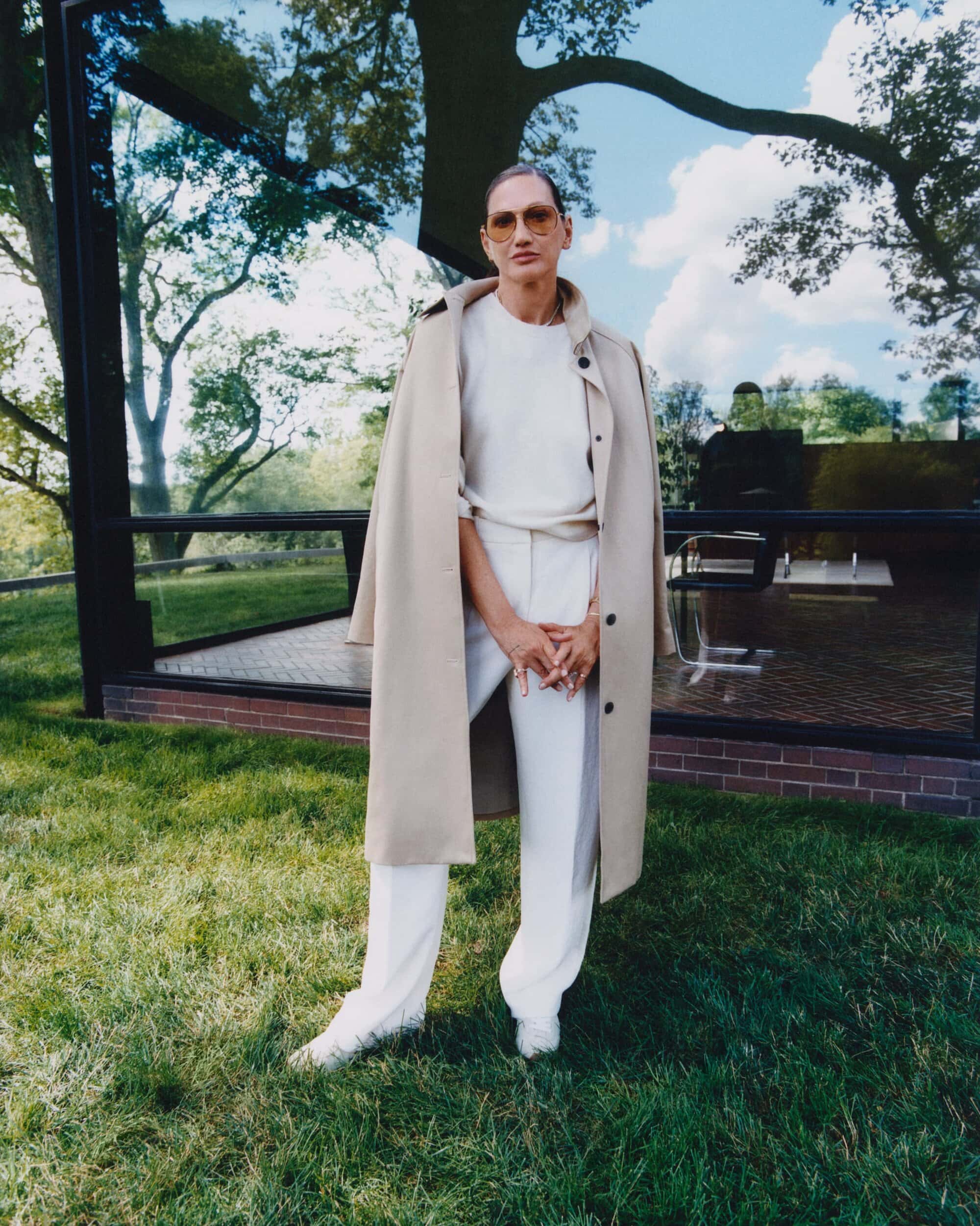Jenna Lyons wearing a beige trench coat and white trousers standing outdoors on a green lawn in front of a modern glass building.