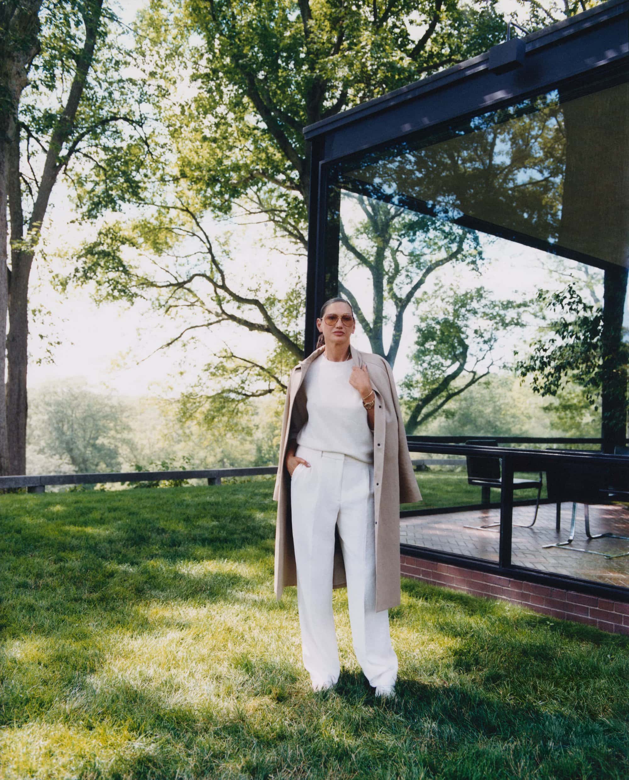 Jenna Lyons standing on a lawn wearing a beige coat draped over her shoulders, white trousers, and sunglasses