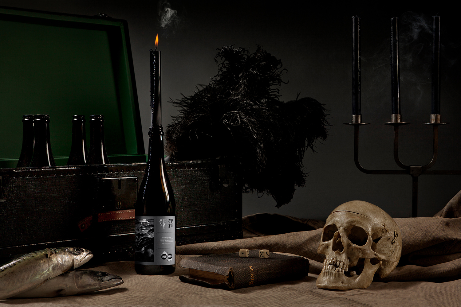 Dark still life with a bottle topped by a lit candle, a human skull, dice, books, fish, and a dark wooden case.