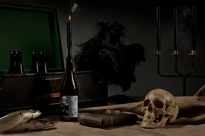 Dark still life with a bottle topped by a lit candle, a human skull, dice, books, fish, and a dark wooden case.