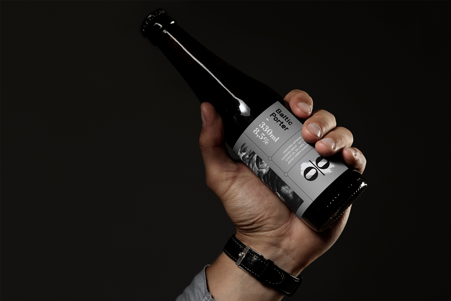 Hand holding a dark glass beer bottle with a grey label against a dark background