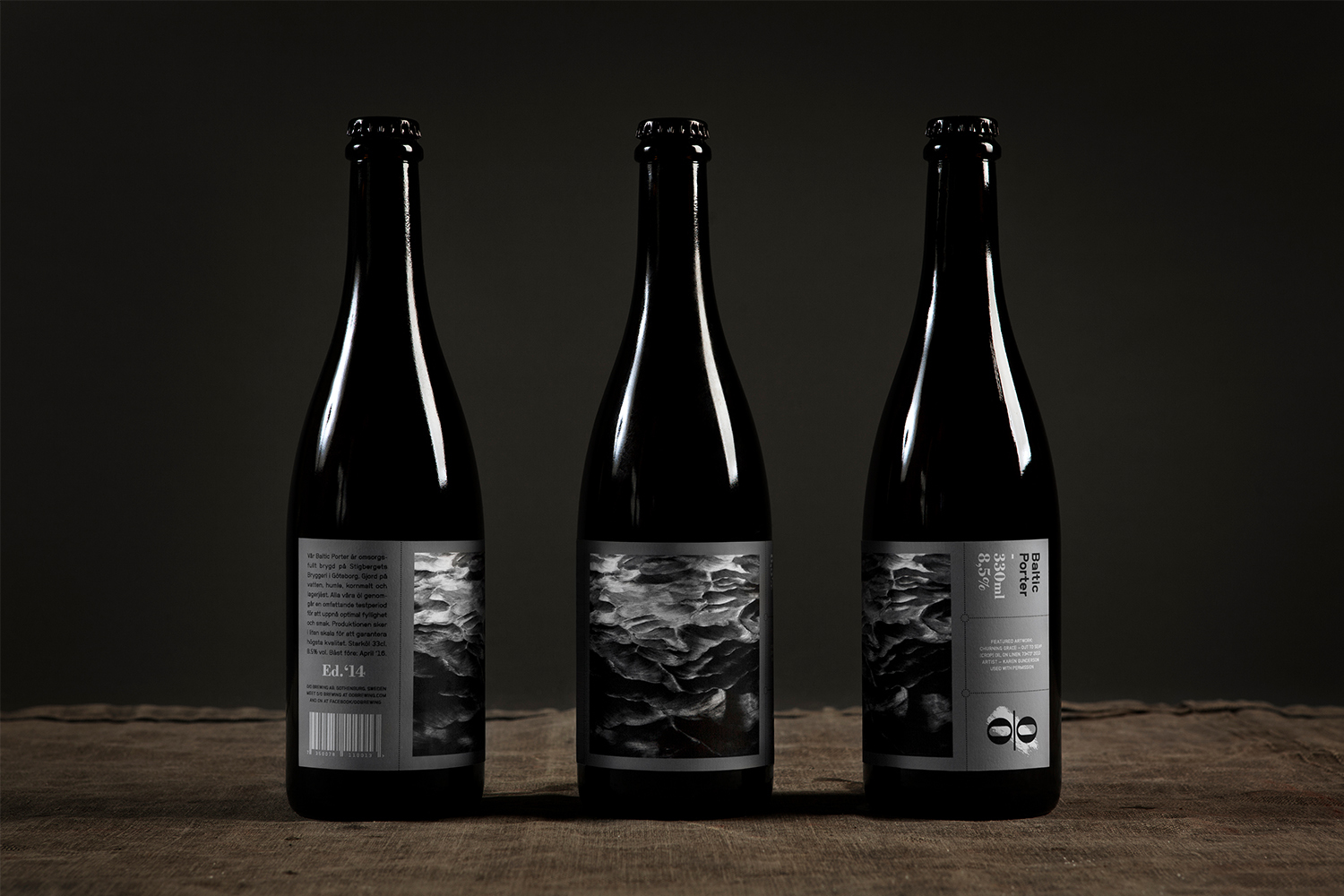 Three dark beer bottles featuring monochrome textured labels arranged upright on a dark textured fabric surface
