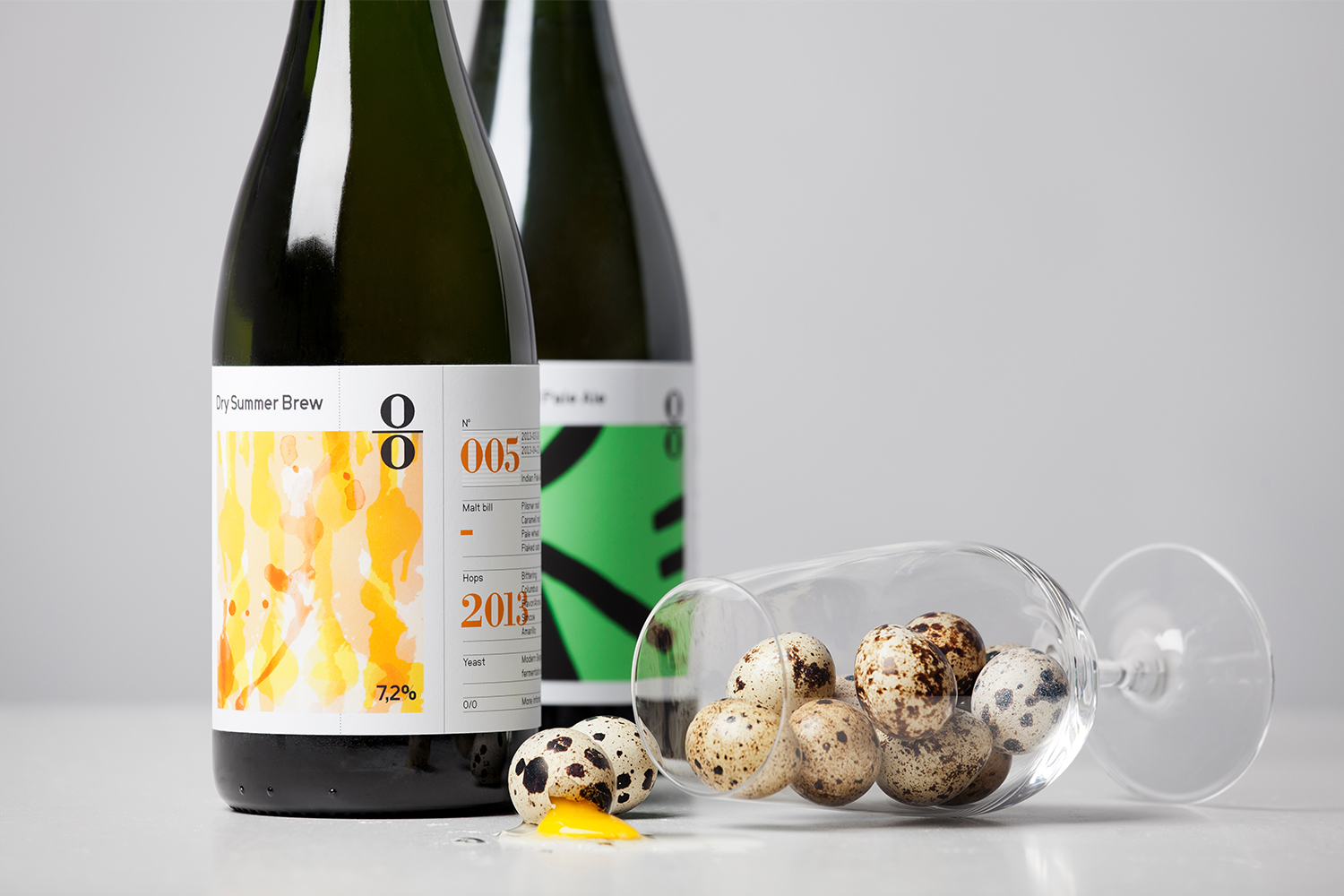 Two O/O Brewing beer bottles standing next to a tipped glass filled with speckled quail eggs and spilled yolk