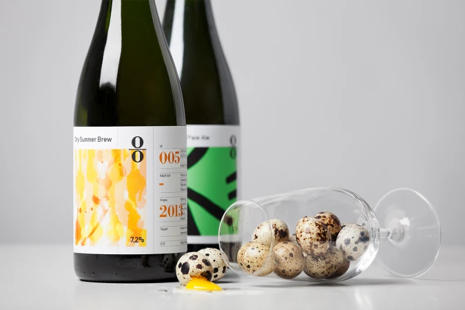 Two O/O Brewing beer bottles standing next to a tipped glass filled with speckled quail eggs and spilled yolk