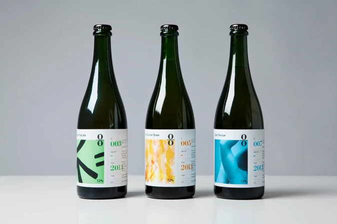 Three dark green glass beer bottles with minimalist white labels stand in a row against a plain grey seamless studio backdrop.